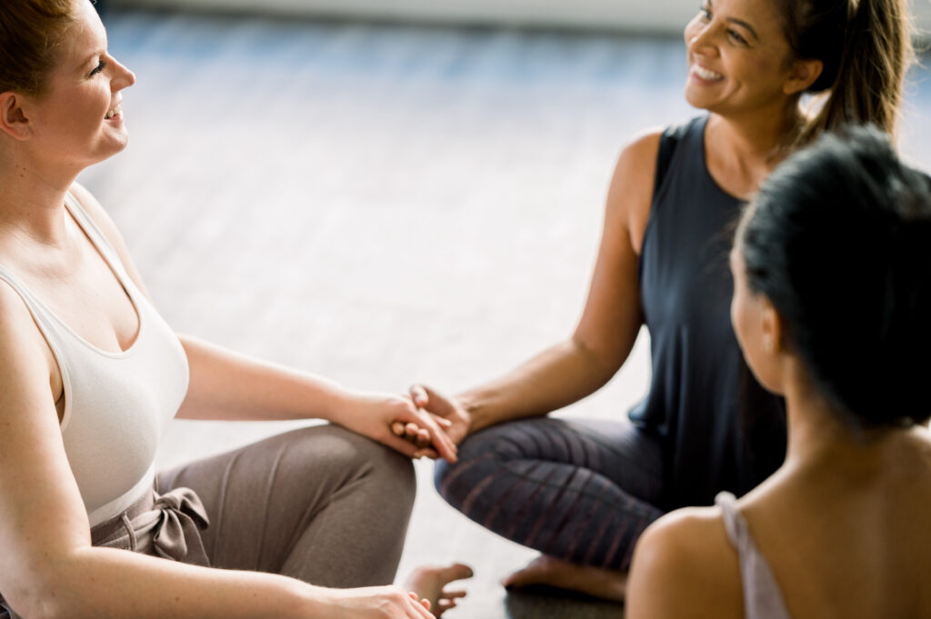 Drei Frauen sitzen im Kreis, halten sich an den Händen und lächeln während einer gemeinsamen Yoga- und Embodiment-Praxis; symbolisiert Verbundenheit, Achtsamkeit und gemeinsames Praktizieren.